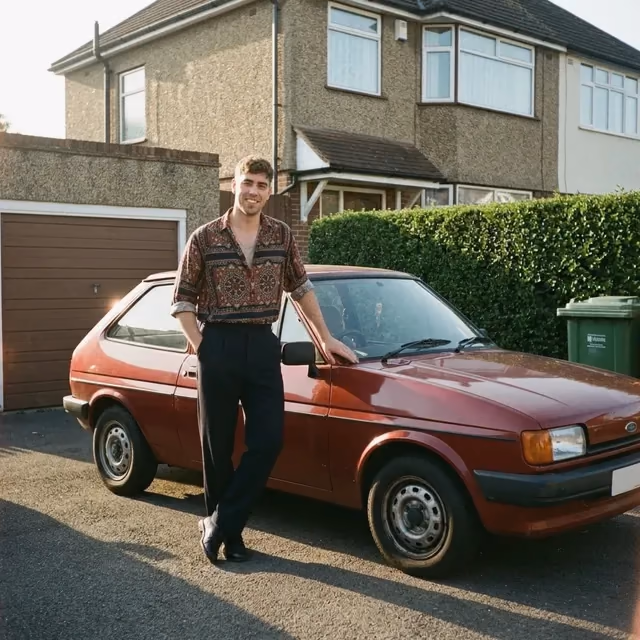 Oliver leaning on a red hatchback on a suburban driveway in the low sun, in a patterned shirt and high-waisted trousers.