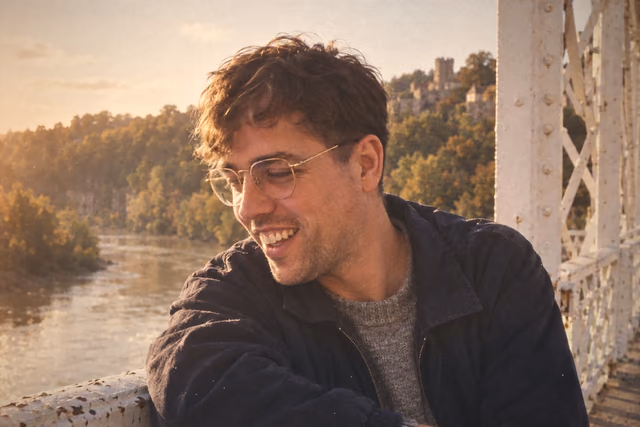 Oliver on an old iron bridge over a river, laughing and looking away downstream, in glasses and a dark jacket, with a castle on the cliff behind him.
